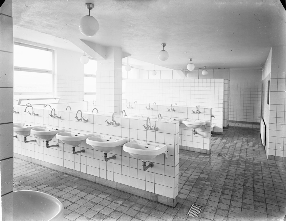 Schwarzes und weißes Foto einer öffentlichen Toilette mit Waschbecken, Wasserhähnen, Wänden, Lampen und Fenstern.