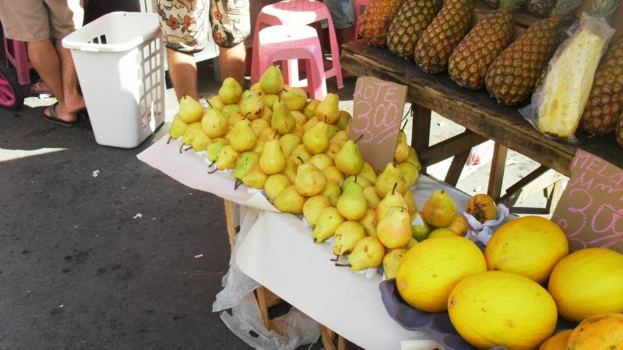 Eine MarktSzene mit verschiedenen Fruchten auf einem Gestell, einigen Hockern, ein paar Menschen und zwei PreisSchildern.