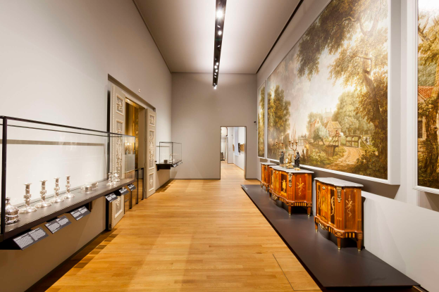 Ein Raum im Stedelijk Museum in Amsterdam, der verschiedene Kunstwerke, darunter Gemälde, Objekte, eine Glasbox mit Gegenständen, Texttafeln und Deckenleuchten zeigt.