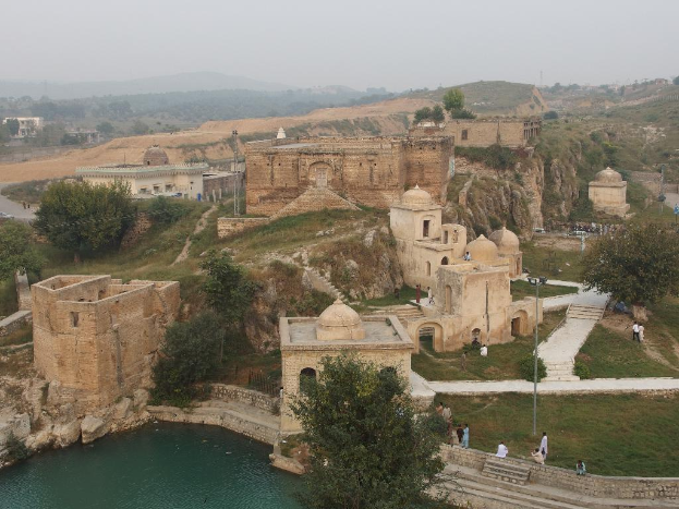 Eine Landschaft mit zahlreichen Burgen und Gebäuden, umgeben von Gras und Bäumen, mit einem Teich im Vordergrund, Hügeln im Hintergrund und einem Himmel darüber.