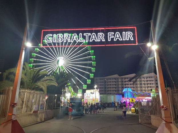 Der Eingang zur Gibraltar-Messe bei Nacht, beleuchtet von einem großen beleuchteten Riesenrad und einer leuchtenden Namensanzeige, mit Menschenmassen drumherum, Bäumen und Gebäuden im Hintergrund unter einem dunklen Himmel.
