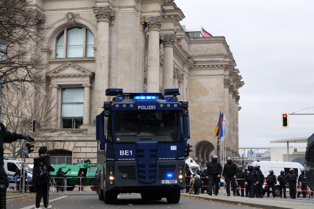 Gruppe von Polizeibeamten vor einem großen Gebäude mit architektonischen Details, Fahrzeugen auf der Straße, einer Person mit einer Kamera auf der linken Seite und Hintergrundelementen wie Bäumen, Ampeln, Fahnen und einem klaren Himmel.