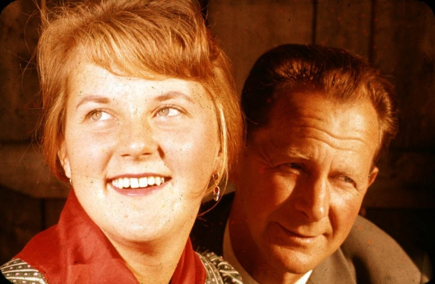 Zwei Menschen umarmen sich eng, lächeln in die Kamera, während der Mann seinen Arm um die Taille der Frau legt und ihr Kopf auf seiner Schulter ruht, vor einer einfachen Wand.