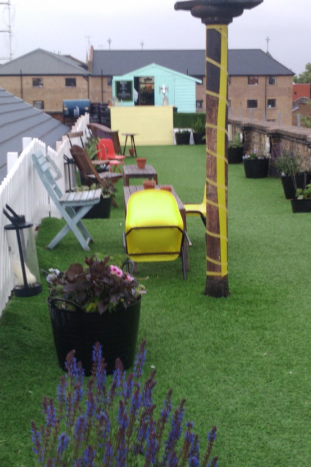 Eine grüne Fläche mit Stühlen, Tischen, Pflanzen und anderen Gegenständen, mit Häusern im Hintergrund und dem Himmel darüber.