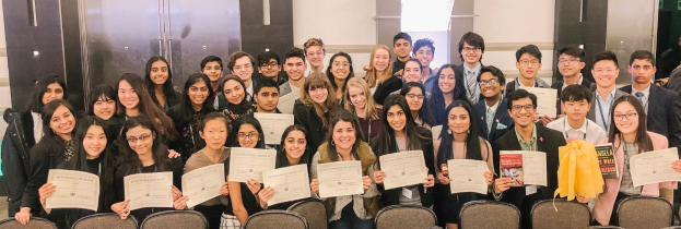 Eine Gruppe von Menschen posiert mit Urkunden vor Stühlen und einer Wand, wahrscheinlich bei einer Abschlussfeier für das akademische Jahr 2019-2020.