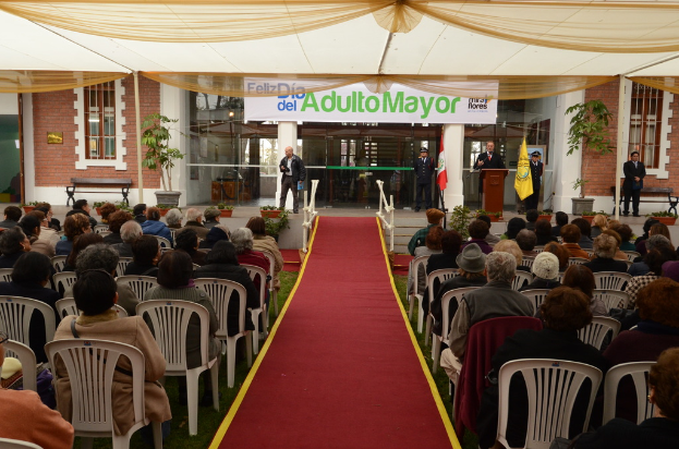 Ein Mann steht vor einem Gebäude mit einem Bogen, auf dem "Adulto Mayor" steht, während im Hintergrund viele Menschen auf Stühlen sitzen.