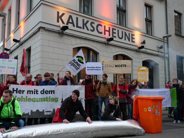 Eine Gruppe von Menschen vor einem Gebäude mit Schildern und Plakaten, zwei Personen im Vordergrund und einem Müllcontainer rechts, während einer Demonstration in Deutschland.