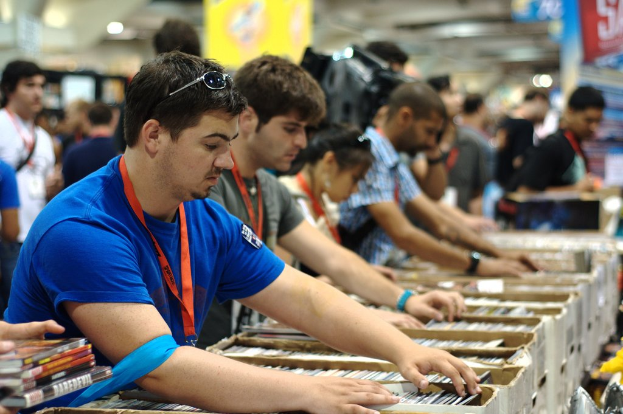 Eine Gruppe von Menschen mit Ausweisen packt Bücher in Kisten, mit Lampen darüber und einem Banner oben rechts, während einige andere im Hintergrund stehen.