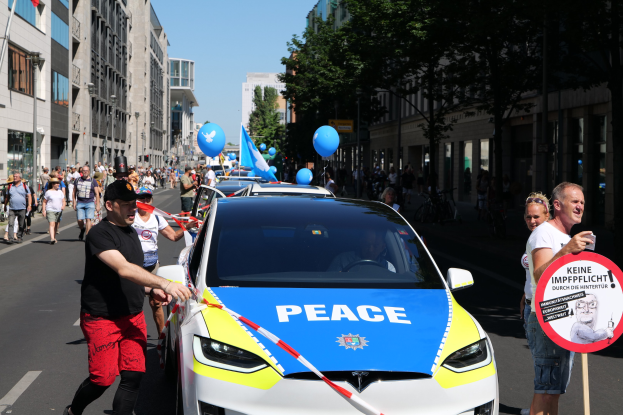 Eine Gruppe von Menschen geht neben einem Polizeiwagen mit blauen, gelben und roten Schleifen und dem Wort "Frieden" darauf, vor einem Hintergrund von Gebäuden, Bäumen und einem klaren Himmel.