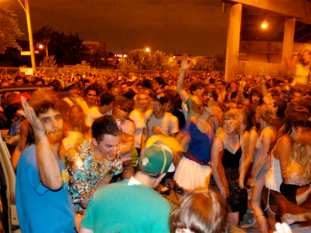 Menschen tanzen im Vordergrund bei einem Konzert mit Lichtern, Polen, Bäumen und anderen Objekten im Hintergrund.