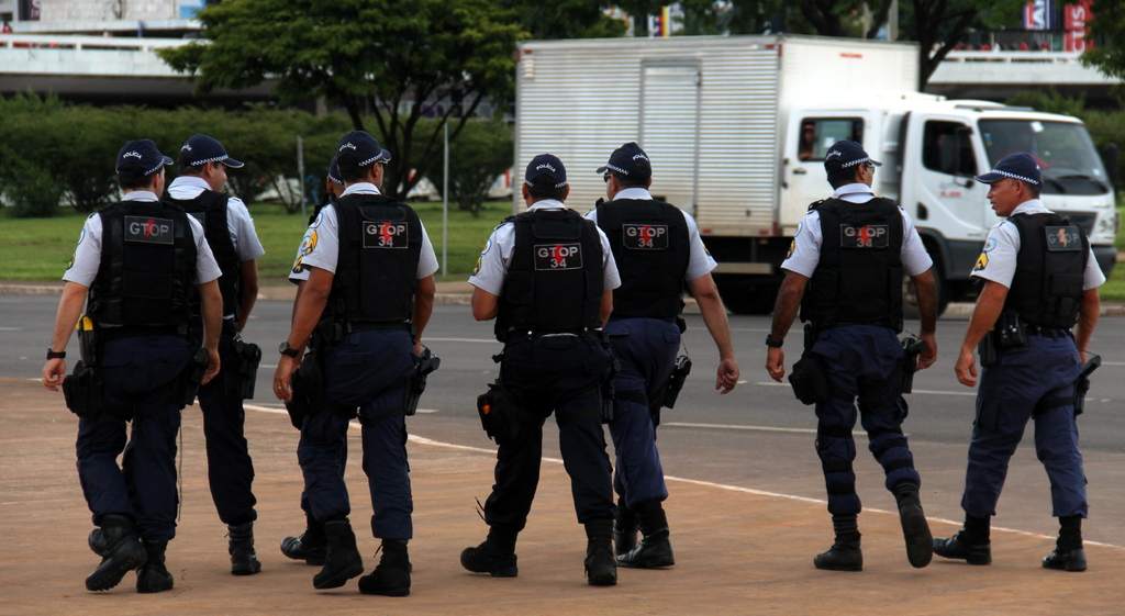 Eine Gruppe von Beamten in Uniform, einschließlich Mützen, Hemden, kugelsicheren Westen, Hosen und Schuhen, geht auf einer Straße. Rechts daneben parkt ein Lastwagen in der Nähe eines Zauns, mit Gebäuden, Bäumen, Pflanzen, Pfählen und Bannern im Hintergrund und einer Brücke oben rechts.