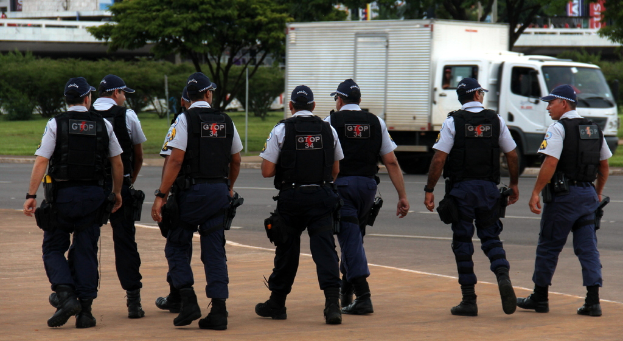 Eine Gruppe von Beamten in Uniform, einschließlich Mützen, Hemden, kugelsicheren Westen, Hosen und Schuhen, geht auf einer Straße. Rechts daneben parkt ein Lastwagen in der Nähe eines Zauns, mit Gebäuden, Bäumen, Pflanzen, Pfählen und Bannern im Hintergrund und einer Brücke oben rechts.