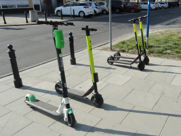 Drei Elektro-Scooter auf einer grasbewachsenen Straßenkreuzung mit Gebäuden, Bäumen und einem klaren Himmel im Hintergrund.