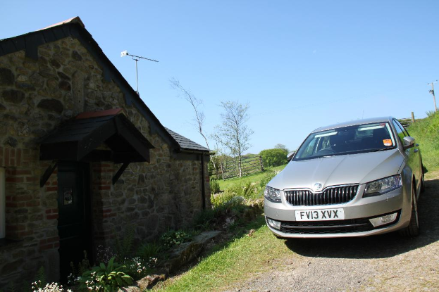 Eine Szene mit einem Haus, einem Zaun und verschiedenen Pflanzen und Bäumen links und einem Auto, einem Strommast und mehr Vegetation rechts unter einem sonnigen Himmel.