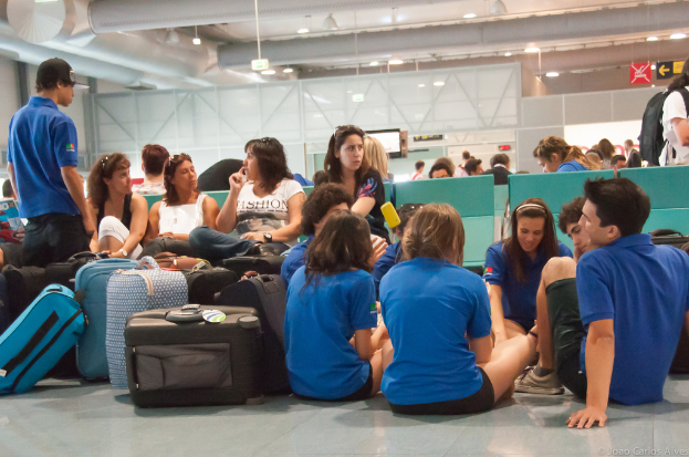 Ein belebter Flughafeninnenraum mit vielen Menschen, die auf ihre Flüge warten, Gepäck auf der linken Seite und gläserne Türen mit Richtungsschildern im Hintergrund.