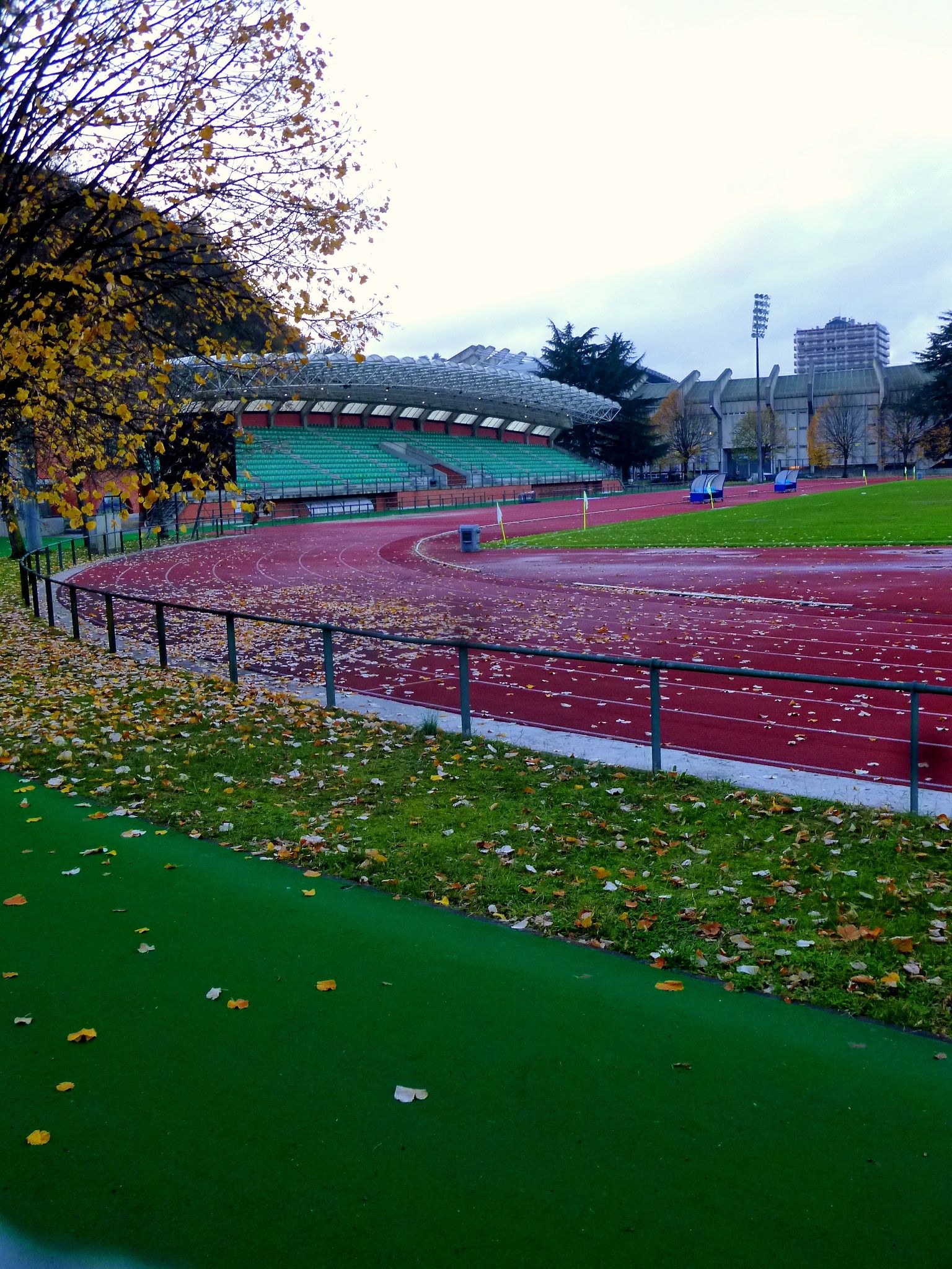 Eine Laufbahn in einem Park mit Herbstlaub auf dem Boden, umgeben von einem Zaun, Gras, Bäumen, Tribünen, Laternenmasten, Gebäuden und einem bewölkten Himmel.