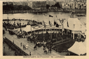 Ein Schwarz-Weiß-Foto einer Menge, die sich um einen Rummelplatz mit Zelten, Fahnen, Stangen und Pferden im Vordergrund und Gebäuden, Bäumen und Wasser im Hintergrund versammelt hat, mit Text am unteren Rand des Bildes.