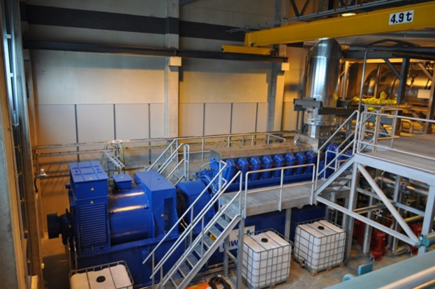 Ein Fabrik-Innenraum mit Trägern, einer Treppe, Geländer, Containern, Lampen, Rohren und Wänden.