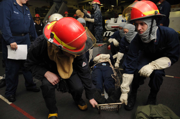 Rettungsteam-Mitglieder in Helmen und Handschuhen helfen einer Person auf einer Trage auf dem Deck eines Schiffs, wobei einer Papiere hält und ein anderer einen Beutel verwaltet.
