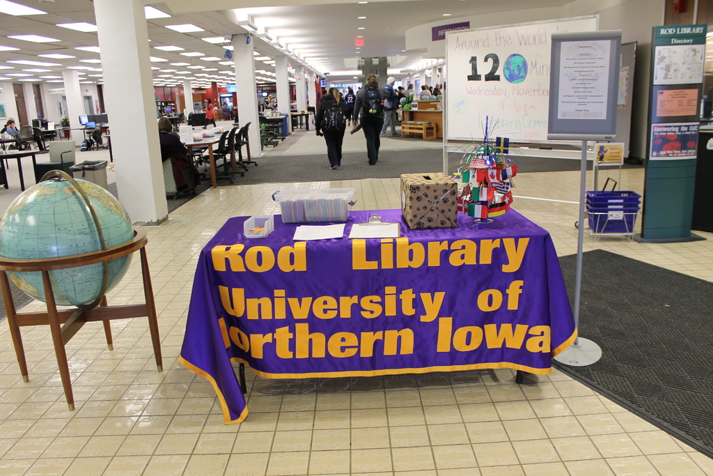 Eine Ausstellung mit einem Tisch, auf dem verschiedene Landesflaggen, eine Box, Papiere und ein Banner liegen, eine Kugel links daneben, ein Anschlagbrett rechts daneben und mehrere Tische und Stühle in der Ferne.