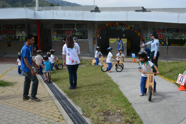 Kinder mit Helmen fahren Fahrräder auf einer Rasenfläche, Erwachsene stehen in der Nähe und ein Schild und ein Gebäude sind im Hintergrund zu sehen.