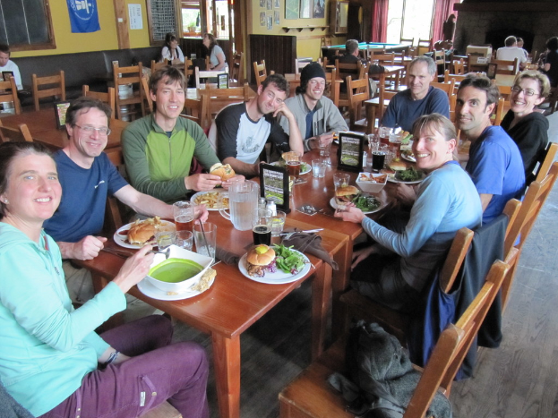 Eine Gruppe von Personen sitzt um einen Tisch mit Tellern, Schüsseln, Gläsern, Flaschen, einem Wasserglas, Karten, Tüchern und Essen, die lächeln, während auf einem der Stühle eine Jacke liegt, und im Hintergrund weitere Personen an anderen Tischen sitzen, mit einer Fahne, Bildern und Plakaten an der Wand und einem Fenster mit Vorhängen.