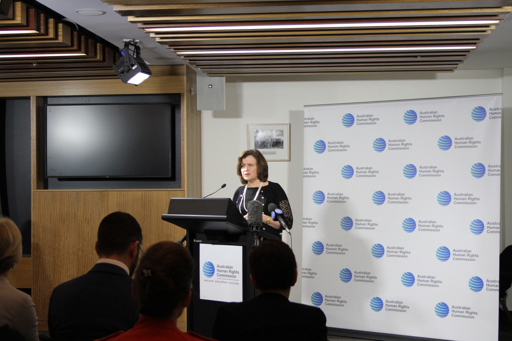 Eine Frau steht auf einer Bühne und spricht in ein Mikrofon, vor ihr sitzen Zuschauer, an der Wand hängt ein Banner, auf dem Holzwand steht ein Fernseher und an der Decke leuchten Lampen.