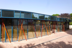 Ein modernes Schulgebäude mit zahlreichen Fenstern, einem baumbestandenen Weg vorne, umgeben von Grün unter einem klaren blauen Himmel.