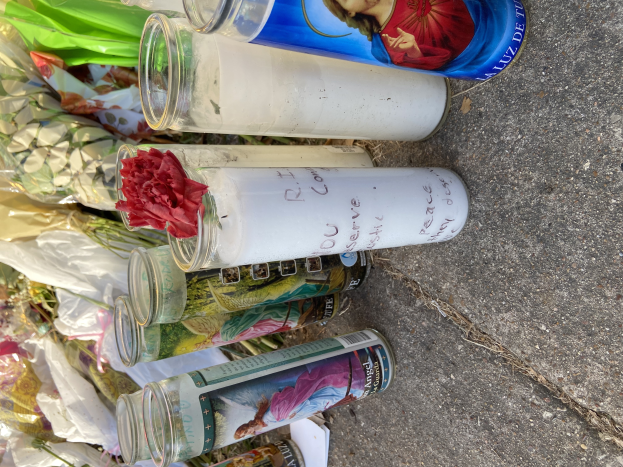 Eine improvisierte Gedenkstätte am Straßenrand mit Glasflaschen, die Fotos und Text zeigen, einem Blumenstrauß und angezündeten Kerzen auf dem Boden.