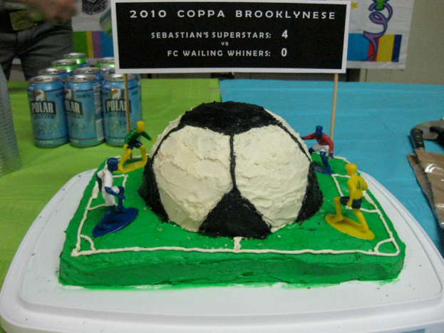 Ein fußballstadionförmiger Kuchen mit einem Fußball und vier Spielern, umgeben von Softdrinkdosen, mit einem Banner im Hintergrund und einer Person oben links, mit einer Tafel im Kuchen.