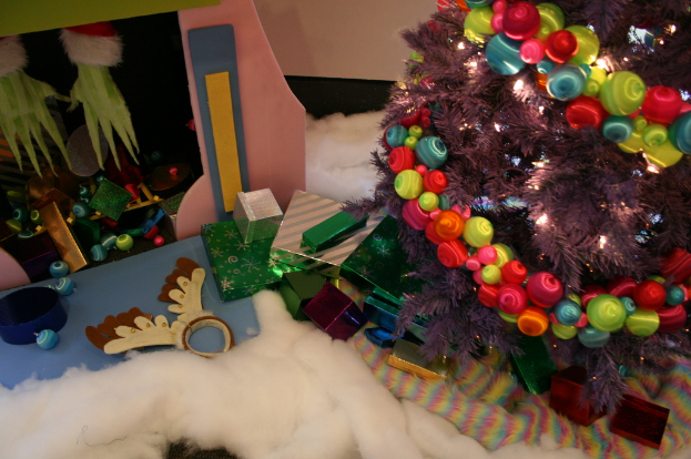 Drinnen steht ein geschm├╝ckter Weihnachtsbaum mit Lichterkette und Schmuck rechts, Geschenkboxen und Deko-Artikel in der Mitte, eine H├╝tte mit Deko oben links und Baumwolle und Wolle unten.