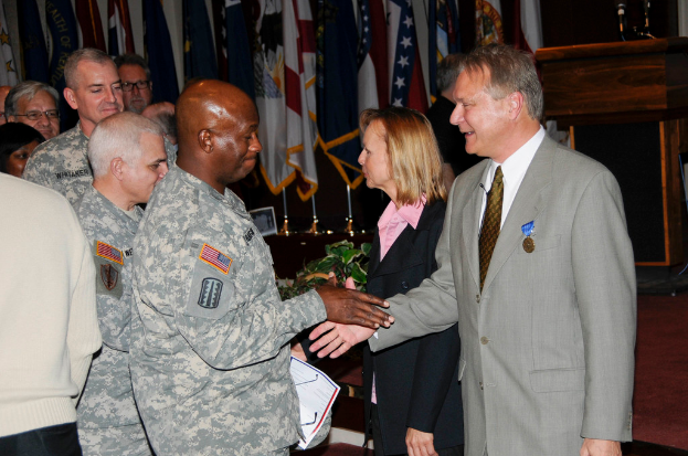 Eine Gruppe von Menschen, die mit zwei Personen Handschlag begrüßen, Flaggen im Hintergrund, ein Podium mit Mikrofon und eine Topfpflanze in der Nähe.