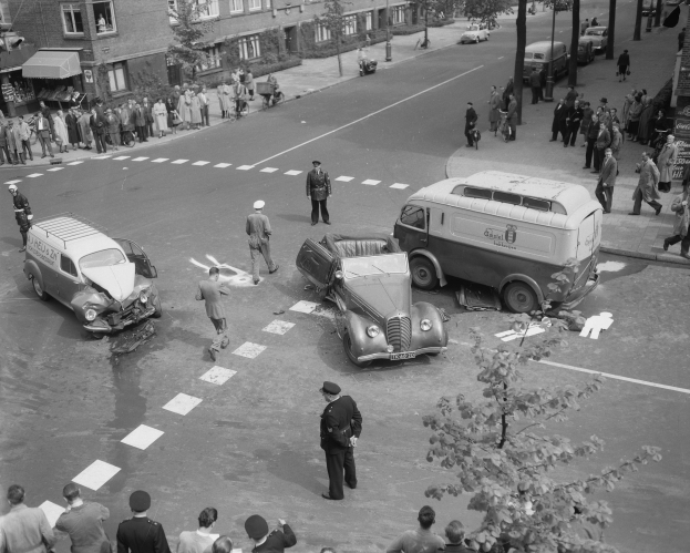 Schwarz-weiß-Szene eines Autounfalls an einer Straßenkreuzung mit mehreren Fahrzeugen, darunter ein Van, und einer Gruppe von Schaulustigen; Gebäude, Bäume, Pfosten und Texttafeln im Hintergrund.