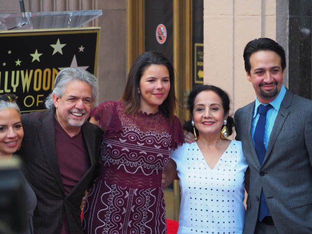 Gruppe von Menschen, die auf einer roten Teppichrolle an der Hollywood Walk of Fame Zeremonie posen, mit einer Kamera auf der linken Seite, einer Tafel mit Text, einem Glaspodest und einer Wand im Hintergrund.