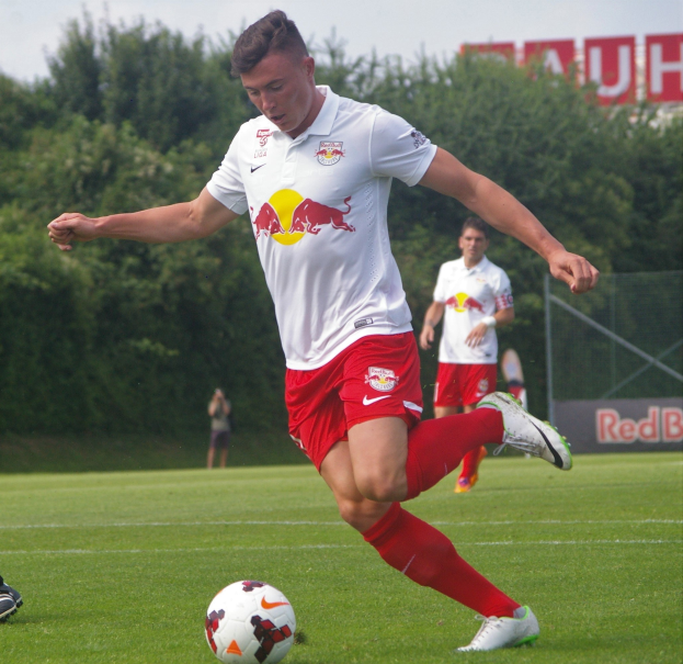 Ein professioneller Fussballspieler in einem roten Trikot schießt einen Ball auf einem grünen Rasen mit Bäumen und einem klaren blauen Himmel im Hintergrund, mit "RB Leipzig" auf einer Tafel dahinter.