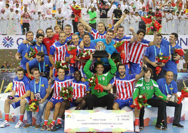 Eine Gruppe von Menschen in Sportkleidung, einige halten Auszeichnungen und Blumen, steht vor einer Menge mit Geländern und Tafeln im Hintergrund.