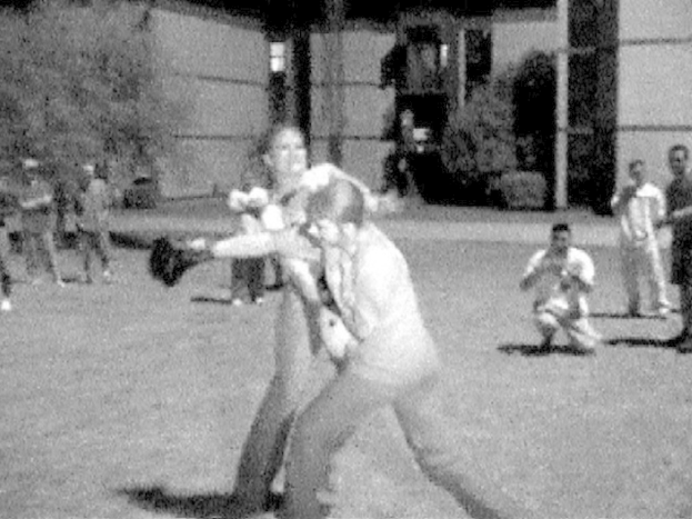 Zwei Personen boxen in der Mitte eines Schwarz-Weiß-Bildes, im Hintergrund ein Gebäude und einige Zuschauer.