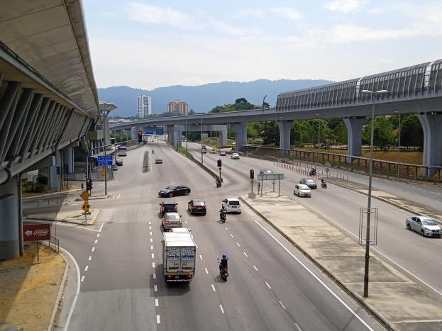 Eine vielbefahrene Autobahn mit zahlreichen Fahrzeugen, umgeben von Straßenlaternen, Verkehrsampeln, Schildern, Bäumen, Gebäuden und Hügeln unter einem bewölkten Himmel.