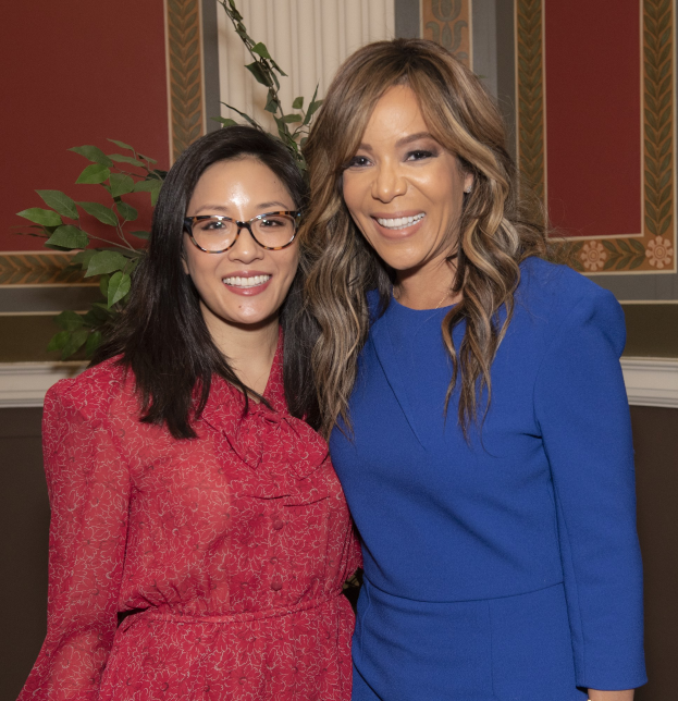 Zwei Frauen, die auf der 2019 Women in Film Pre-Oscar-Lunch-Veranstaltung zusammen lächeln, eine in einem blauen Kleid und die andere in einem roten Kleid mit Brille, stehen neben einer Pflanze und einem Vorhang.