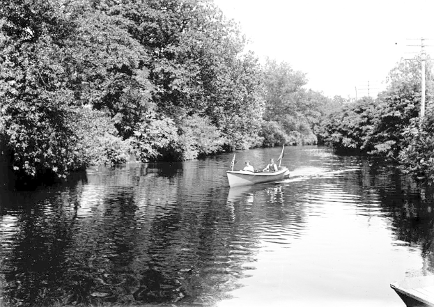 Ein Boot mit Menschen und Flaggen auf dem Wasser, mit BÃ¤umen und MÃ¤sten im Hintergrund.