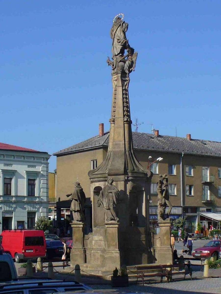 Ein Denkmal steht in einem Stadtplatz umgeben von Gebäuden, Straßenmöbeln, Fahrzeugen, Fußgängern, Bänken, Pflanzen und einer bewölkten Himmel.