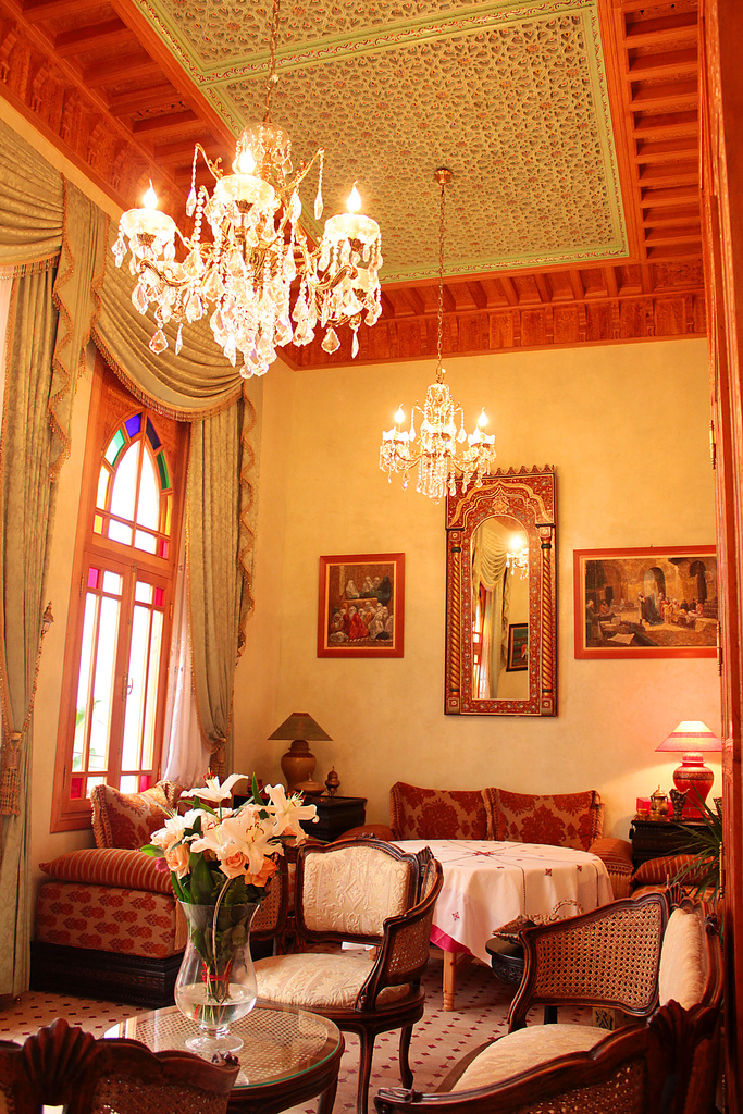 Ein Wohnzimmer mit einem Tisch und Blumentopf im Vordergrund, umgeben von Stühlen, Sofas und einem mit einer weißen Decke bedeckten Tisch im Hintergrund, gerahmte Bilder an der Wand, von der Decke hängende Lampen und Vorhänge mit einem Fenster links.