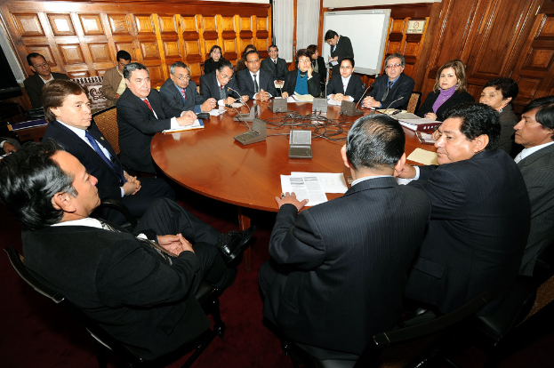 Eine Runde-Tisch-Sitzung mit Delegierten, die diskutieren, Mikrofone, Papiere, Kabel und Dateien auf dem Tisch und eine hölzerne Wand im Hintergrund.