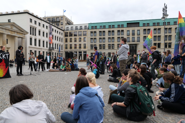 Menschen, die auf dem Boden vor einer Menge sitzen, die Fahnen und Transparente hölt, bei einer Demonstration in Berlin mit einem Mikrofonständer, einer Statue und Gebäuden im Hintergrund.