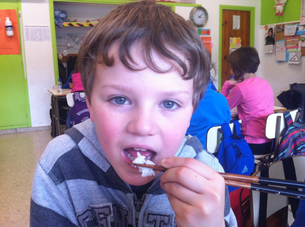 Ein Junge in einer grauen und schwarzen Jacke isst mit Stäbchen in einem Klassenzimmer, mit anderen Kindern im Hintergrund, einer grün-weißen Wand, Bildtafeln an einer weißen Wand, einer braunen Tür und einer Wanduhr.