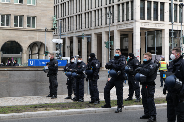 Gruppe von Polizisten in schwarzen Uniformen und Masken vor einem Berliner Gebäude mit Glasfenstern und Säulen, die Helme halten, mit Laternen, Verkehrszeichen, Schildern mit Text und einer Statue im Hintergrund.