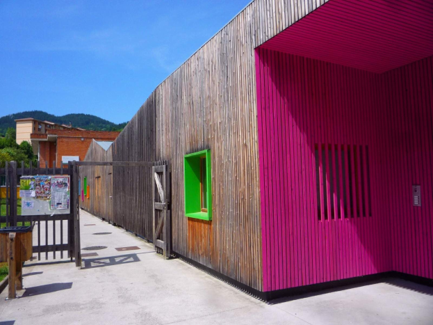 Ein rosafarbenes Geb├Ąude mit einer grünen Tür und Fenstern, einem h├Âlzernen Zaun, einer Tafel mit Papieren, einem Müllcontainer, Gras, B├Ąumen, entfernten Geb├Ąuden, Hügeln und einem bew├Âlktem Himmel, Teil eines umgestalteten Schulspielplatzes in Spanien.