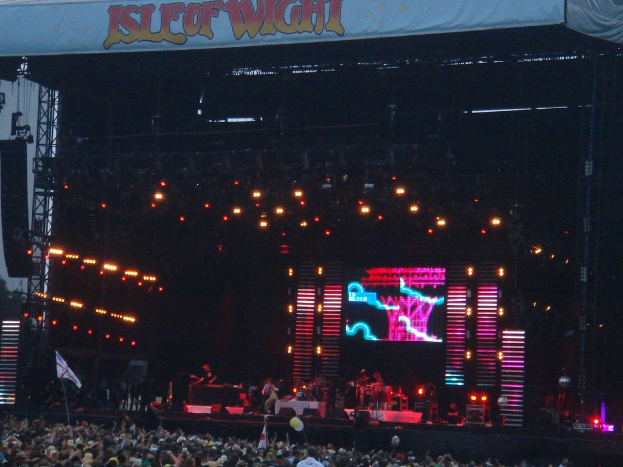 Eine Bühne mit Lichtern, einem Bildschirm und Musikern mit Instrumenten, mit einem Banner oben, einer Menge unten und Fahnen mit Stangen und Ballons.