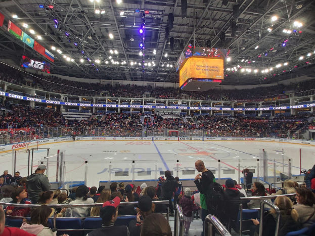 Eine große Menschenmenge in einer Hockeyarena, mit einigen Sitzenden und anderen Stehenden, die ein Spiel unter hellen Scheinwerfern und Bannern beobachten.