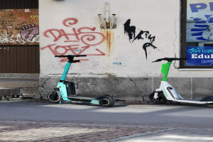 Zwei Elektro-Scooter nebeneinander auf einer Straße geparkt, mit einer Graffiti-bedeckten Wand und einem Plakat an einem Fenster im Hintergrund.